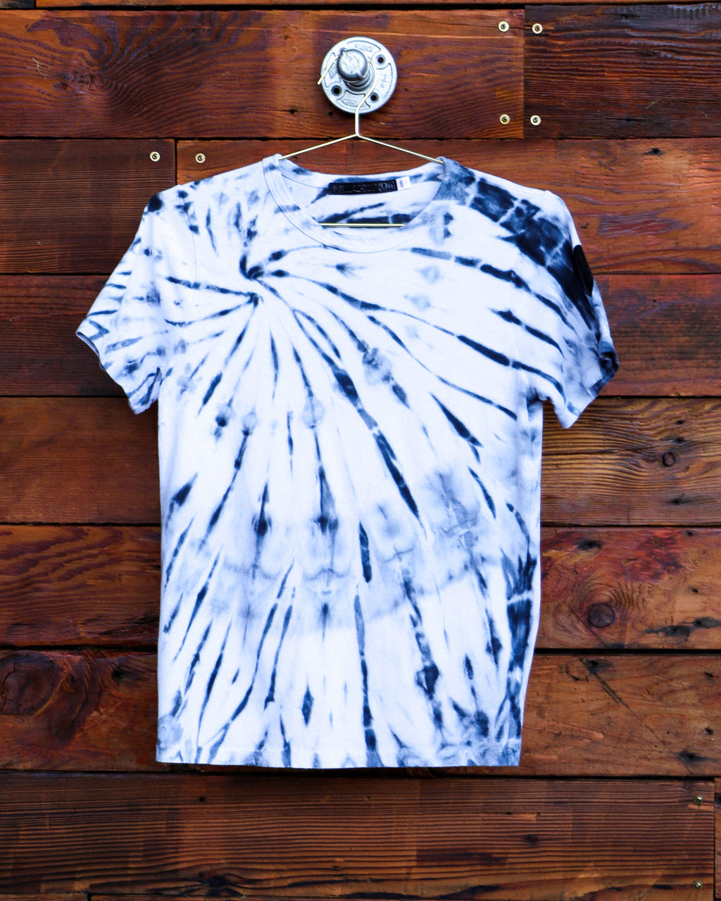 White hand dyed spiral tie dye t-shirt on a hanger in front of a weathered wood wall